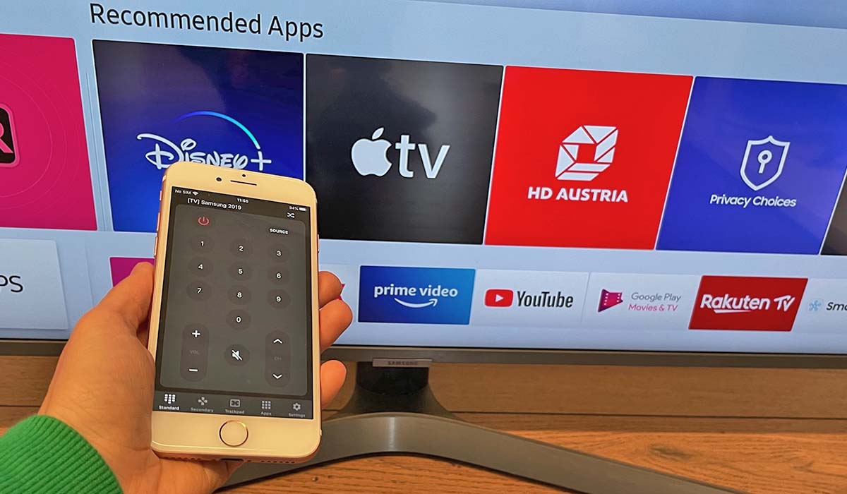 A hand holding a white iPhone with the ControlMeister interface on the screen. A Samsung Smart TV with app icons on the screen