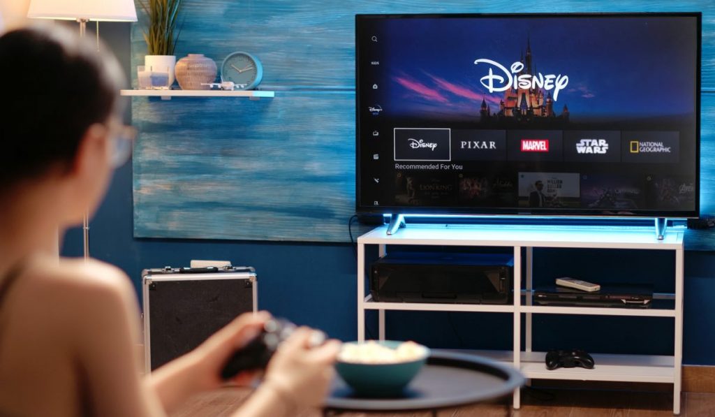 A woman sitting with a controller in her hands. In front of her is a coffee table with a bowl of popcorn on it. A Smart TV with the Disney Plus main menu on the screen